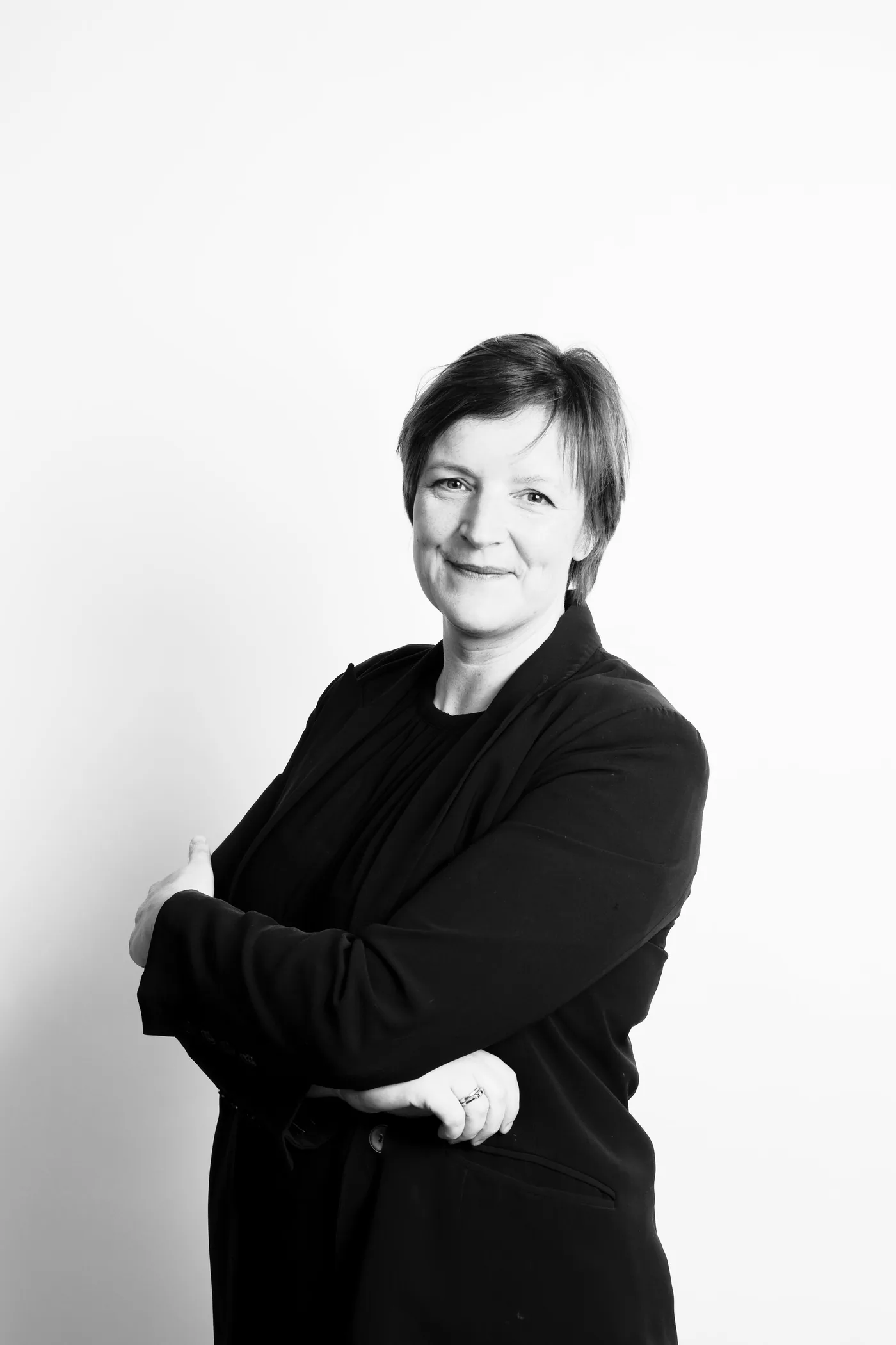 Zwarte en witte portretfoto van een vrouw met kort haar die lacht, gekleed in zwarte kleding, met de armen over elkaar, tegen een witte achtergrond.